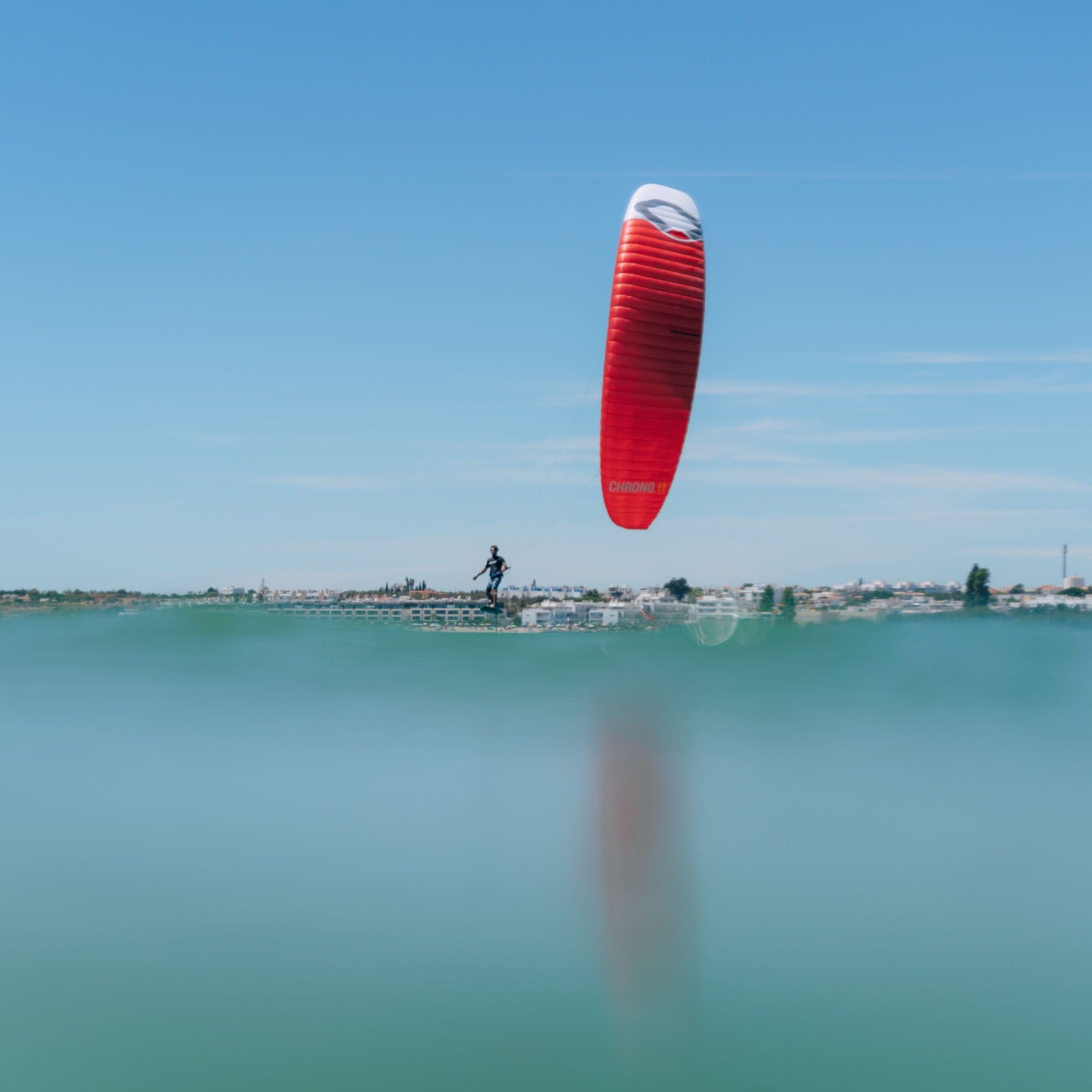 A kitesurfer flying a red Ozone Chrono V4 on mirror flat blue water.