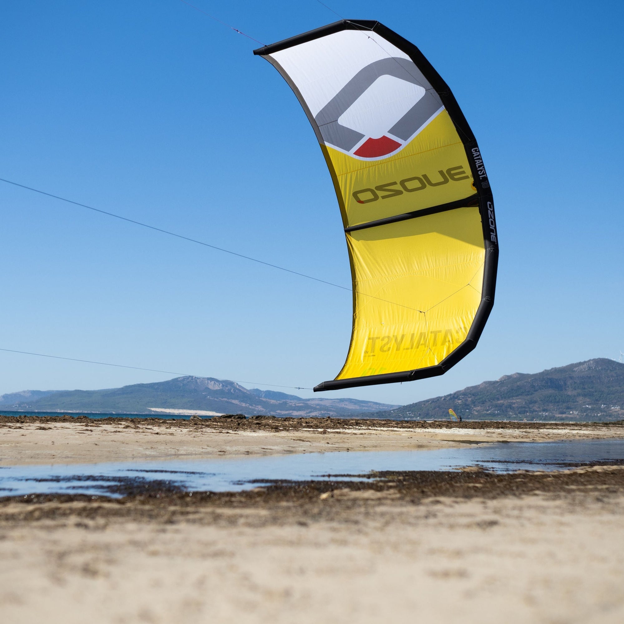 The Ozone Catalyst V3 in yellow flying above a beach in clear blue sky.