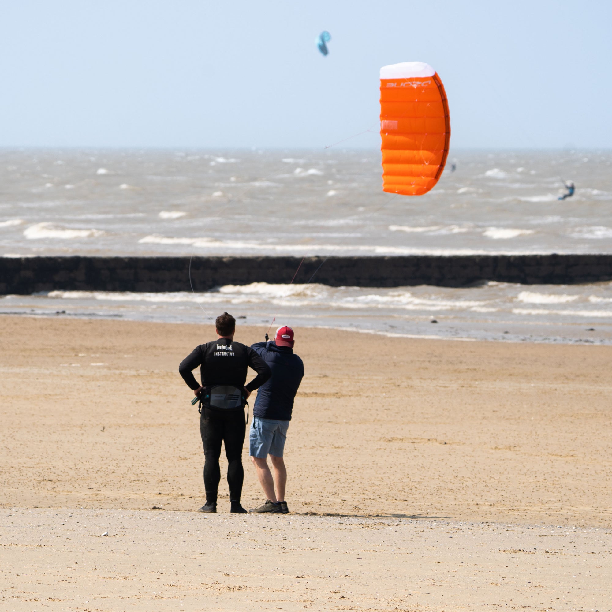 Kitesurfing taster lesson in progress on the golden sand beach of Minnis Bay near Margate, Kent. The instructor is flying an orange Ozone Ignition trainer kite to show them how to fly it in the wind window.